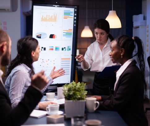 Multi-ethnic businesspeople discussing financial company solution sitting at conference table in meeting room. Diverse teamwork overworking at management strategy using tv late at night.